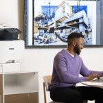 Xerox® VersaLink® C415 Farb-Multifunktionsdrucker in einem Büro Männer arbeiten an einem Laptop vor einem Xerox® VersaLink® C415 Farb-Multifunktionsdrucker