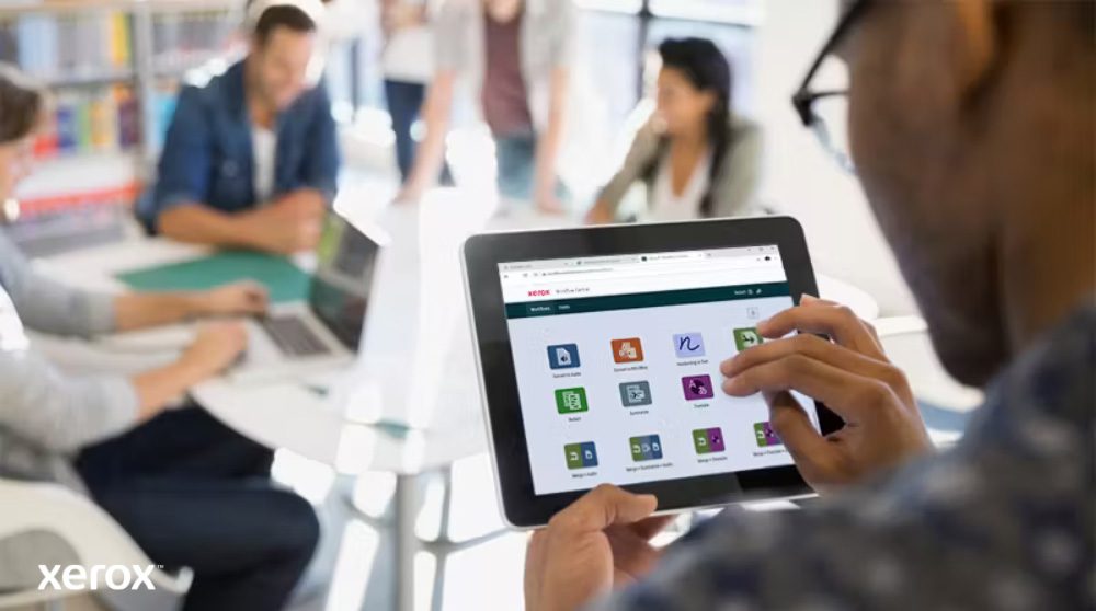 Xerox® Workflow Central Platform: Intuitiver mobiler Zugriff Eine Person hält ein Tablet, auf dem die Benutzeroberfläche von Xerox Workflow Central mit verschiedenen App-Symbolen angezeigt wird, während andere Personen im unscharfen Hintergrund arbeiten und den mobilen Zugriff auf Workflow-Lösungen hervorheben.