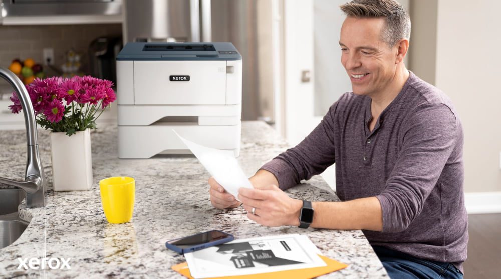 "Xerox® Druck- und Scan-Erlebnis: Mühelose Dokumentenverwaltung " Eine Person lächelt, während sie an einer Küchentheke ein Dokument überprüft. In der Nähe stehen ein kompakter Xerox-Drucker und eine leuchtend gelbe Tasse, was auf nahtloses Drucken und Dokumentenhandling zu Hause oder in einem kleinen Büro hindeutet.