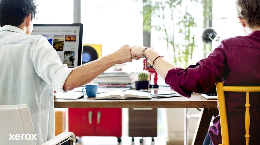 Zwei Personen sitzen an einem Schreibtisch und blicken von der Kamera ab. Sie stoßen sich mit den Fäusten aneinander, um Teamwork und erfolgreiche Zusammenarbeit zu symbolisieren. Eine Person blickt auf einen Computerbildschirm.