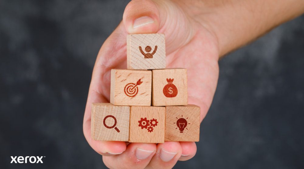 Eine Hand hält einen Stapel Holzklötze, in die jeweils ein Geschäftssymbol eingraviert ist: eine Person, ein Ziel, ein Geldbeutel, eine Lupe, Zahnräder und eine Glühbirne, die verschiedene Vorteile und Lösungen darstellen.
