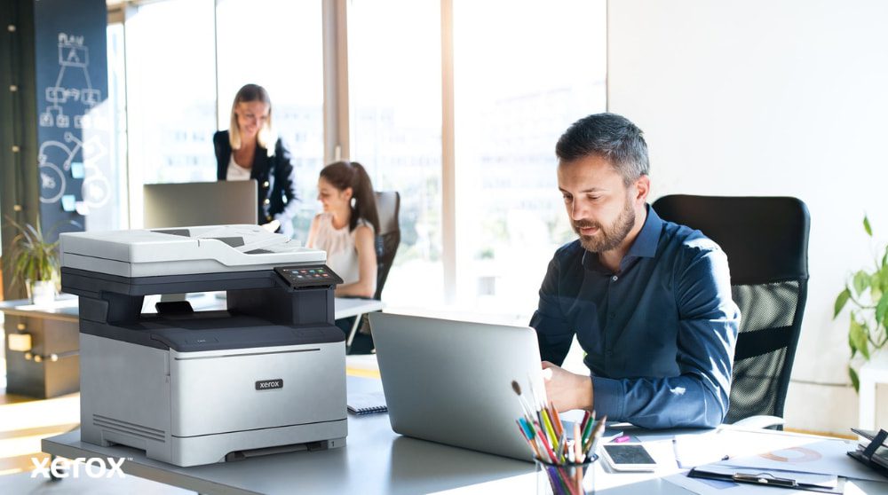 Eine Person sitzt an einem Schreibtisch und blickt auf einen Laptop, daneben steht ein Xerox C325 Multifunktionsdrucker. Im Hintergrund arbeiten zwei weitere Personen ebenfalls an ihren Schreibtischen in einer hellen Büroumgebung.