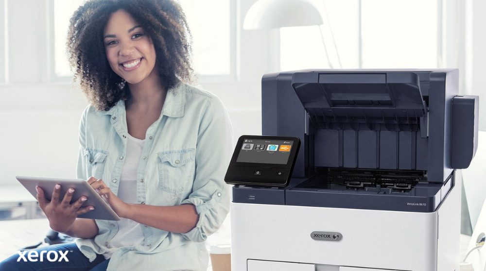 Eine lächelnde Person sitzt neben einem weißen Xerox VersaLink B610-Drucker, hält ein Tablet und blickt in die Kamera, was die Benutzerfreundlichkeit der Xerox-Lösungen hervorhebt.
