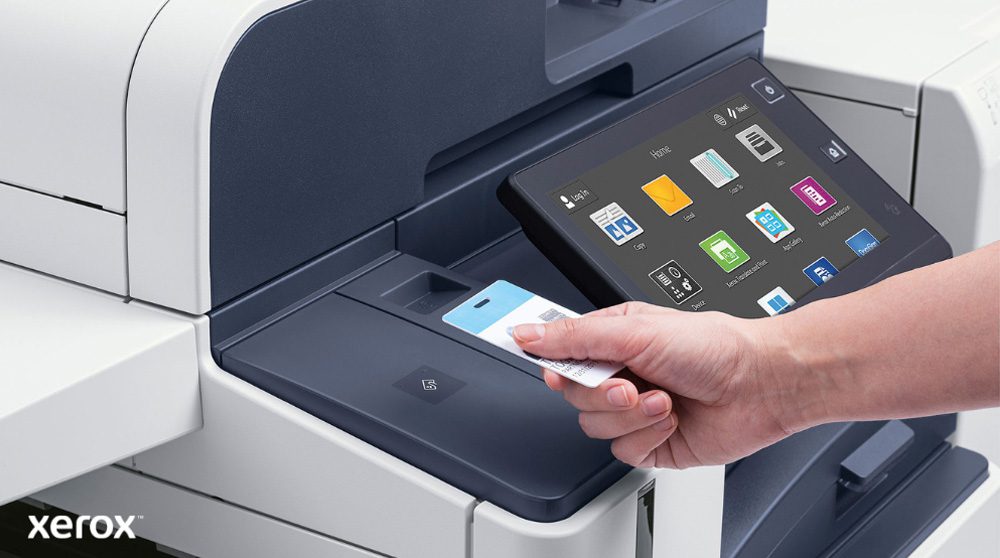 Eine Hand hält eine Sicherheitskarte und scannt sie in der Nähe des Kartenlesers eines modernen Xerox-Multifunktionsdruckers mit einem großen Touchscreen-Display, auf dem verschiedene Anwendungssymbole angezeigt werden.